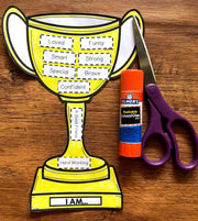 Yellow trophy-shaped paper craft with adhesive labels, Elmer's glue stick, and scissors on a wooden surface.