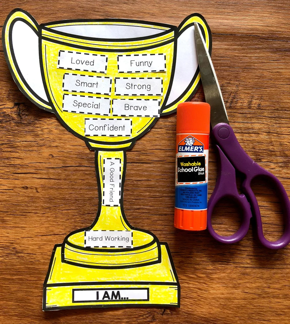 Yellow trophy-shaped paper craft with adhesive labels, Elmer's glue stick, and scissors on a wooden surface.